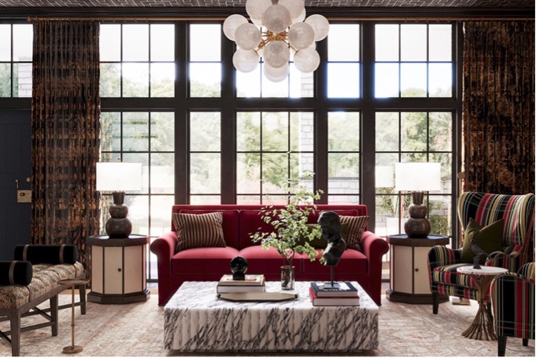 Elegant living room with floor-to-ceiling black-framed windows behind a red velvet sofa, flanked by matching lamps on round side tables, a marble coffee table, and a striped accent chair.