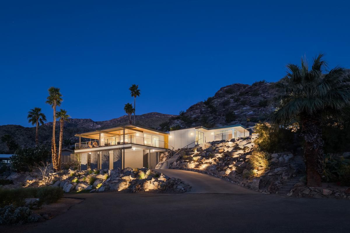 A modern home built into a hillside is illuminated at night, and the lights show off the rocky landscape and the home, which has floor-to-ceiling glass windows and doors. 