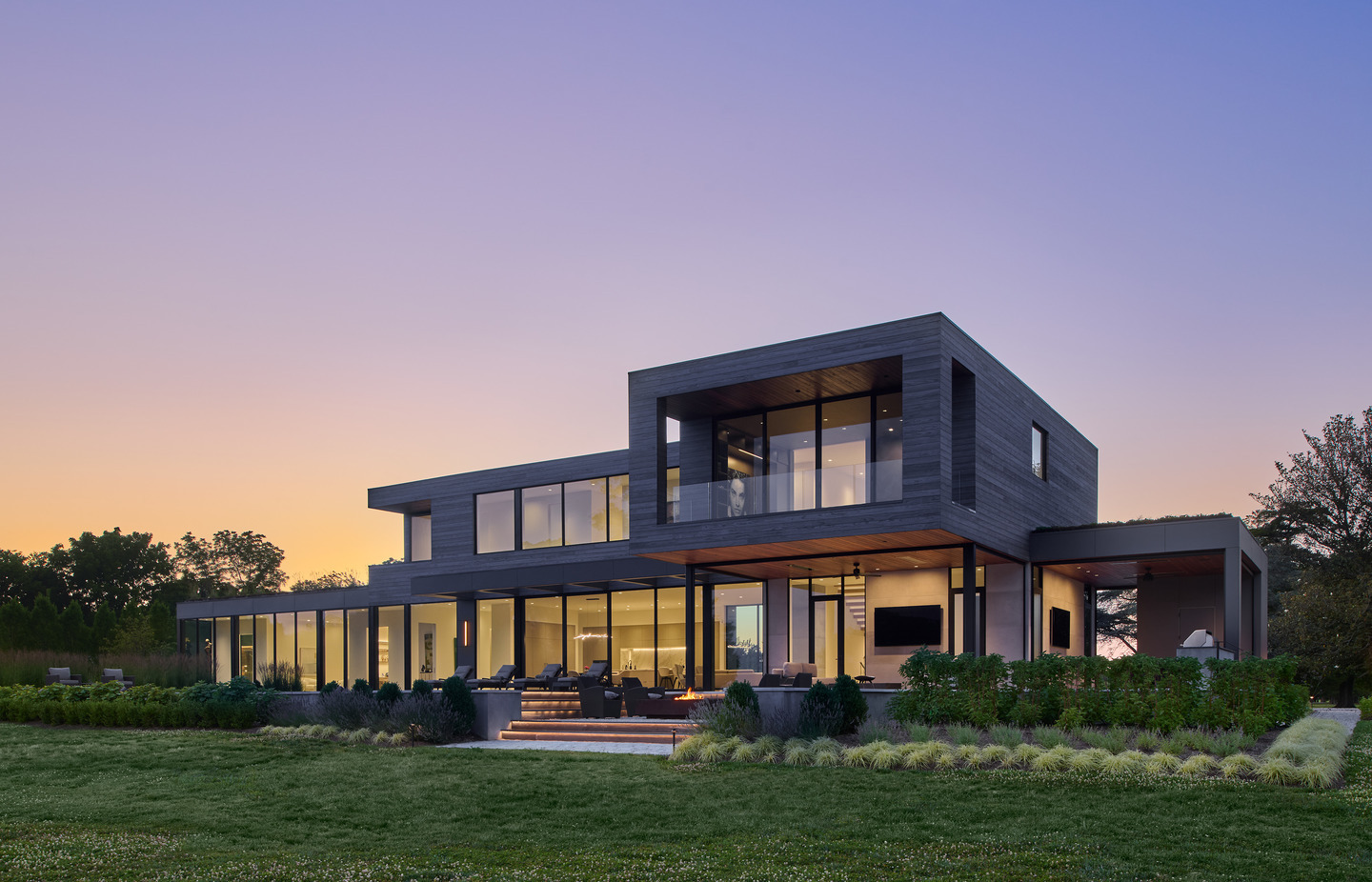 Modern two-story home featuring expansive floor-to-ceiling windows and doors overlooking a landscaped backyard at sunset.