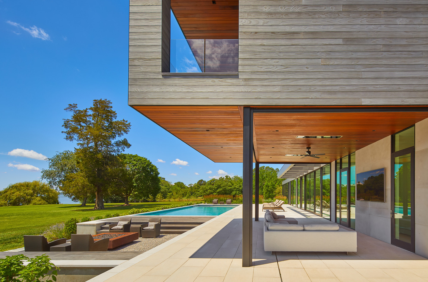 Covered outdoor living area with floor-to-ceiling glass doors overlooking a pool and waterfront lawn.