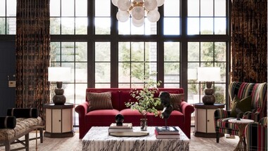 Elegant living room with floor-to-ceiling black-framed windows behind a red velvet sofa, flanked by matching lamps on round side tables, a marble coffee table, and a striped accent chair.