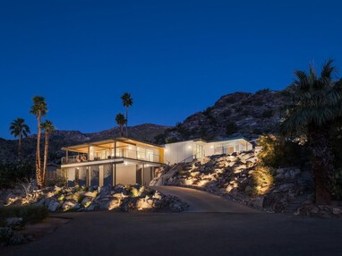 A modern home built into a hillside is illuminated at night, and the lights show off the rocky landscape and the home, which has floor-to-ceiling glass windows and doors. 