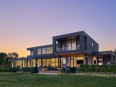 Modern two-story home featuring expansive floor-to-ceiling windows and doors overlooking a landscaped backyard at sunset.