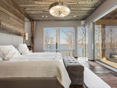 This master bedroom has a massive sliding glass door that leads to a private balcony next to windows with views of a lake.
