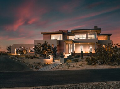 A front view of the glassy, two story home in the desert during sunset.