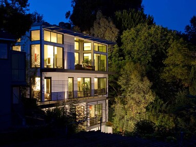 A nighttime view of the three-story home with large doors and panels of glass that lead to the tree-laden landscape.