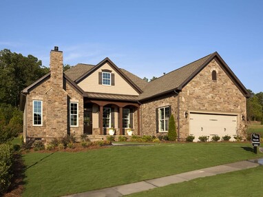 A view of the outside of a one-story house with warm brown brick framed by bright green trees and grass.