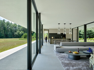A figure is seen walking down the passage of a glass-walled home. The glass wall makes the surrounding field visible.