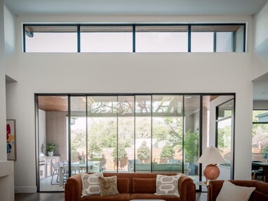 A modern interior of a residential living room shows a sofa in the foreground with a large multi-sliding door with clerestory windows above it. The backyard can be seen just outside. The whole room seems to glow from the light coming from the outside.