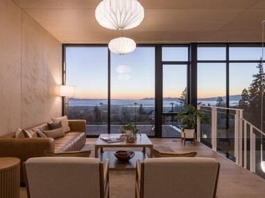 A loft in a modern home has a living room setup, with the staircase going down on the right. The sunset can be seen across a bay through the floor-to-ceiling windows that make up the entire far wall.