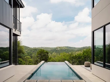 The pool, surrounded by glazing from u-shaped home, overlooks green forestry.
