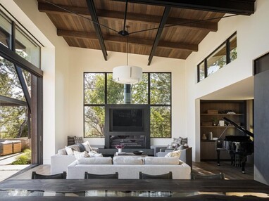 A living room with high wooden ceilings is filled with natural light from the open sliding glass door that makes up most of the wall to the left, a window wall surrounding a large TV directly opposite the viewer, and clerestory windows above the connecting room to the right.