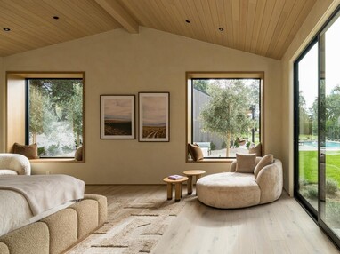 A bedroom with large fixed windows and a sliding door is filled with natural light, warm wood tones, and comfortable seating.