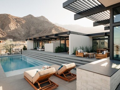The back of a modern desert home has multiple floor-to-ceiling sliding glass doors that open onto a patio with steps that lead down to a pool. Mountains and foothills create the dramatic setting ever-changing backdrop.