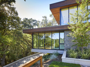 Two stories of a modern home with entirely glass walls reflect the trees across the driveway.