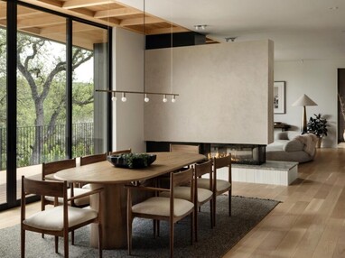 A modern dining room with natural accents leads to a balcony through a floor-to-ceiling multi-slide door. The balcony overlooks trees and a lake is visible through them.