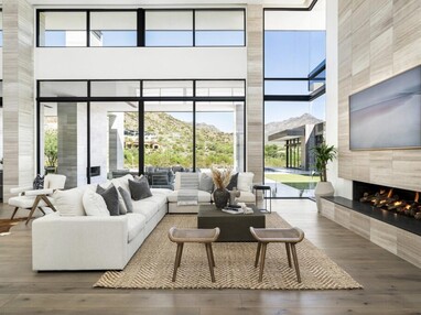 A contemporary living room is set against large glass sliding doors and windows that look out to the backyard and beyond to distant neighbors’ homes, and mountains beyond that.
