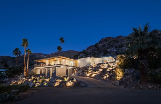 A modern home built into a hillside is illuminated at night, and the lights show off the rocky landscape and the home, which has floor-to-ceiling glass windows and doors. 