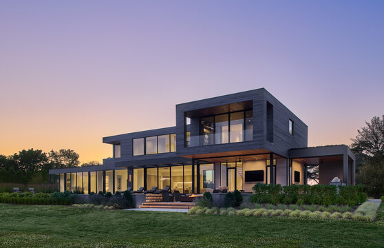 Modern two-story home featuring expansive floor-to-ceiling windows and doors overlooking a landscaped backyard at sunset.