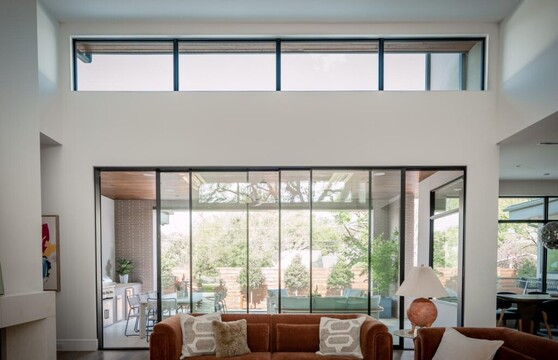 A modern interior of a residential living room shows a sofa in the foreground with a large multi-sliding door with clerestory windows above it. The backyard can be seen just outside. The whole room seems to glow from the light coming from the outside.