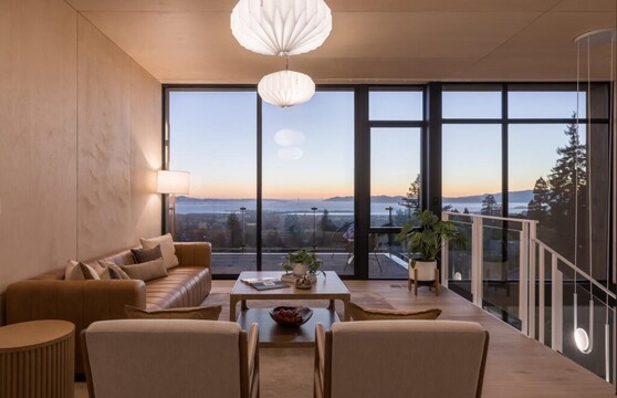 A loft in a modern home has a living room setup, with the staircase going down on the right. The sunset can be seen across a bay through the floor-to-ceiling windows that make up the entire far wall.