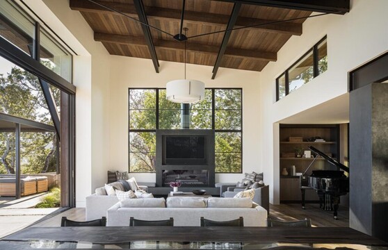 A living room with high wooden ceilings is filled with natural light from the open sliding glass door that makes up most of the wall to the left, a window wall surrounding a large TV directly opposite the viewer, and clerestory windows above the connecting room to the right.