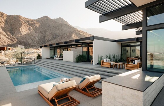 The back of a modern desert home has multiple floor-to-ceiling sliding glass doors that open onto a patio with steps that lead down to a pool. Mountains and foothills create the dramatic setting ever-changing backdrop.