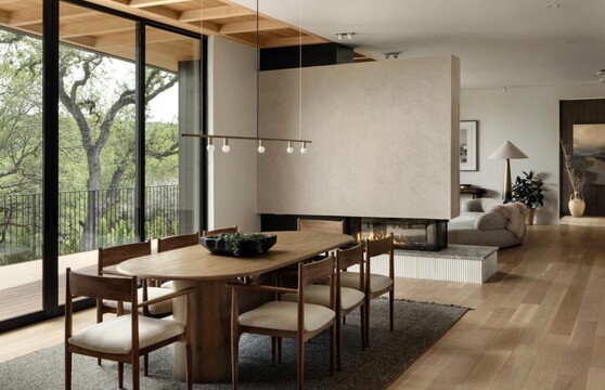 A modern dining room with natural accents leads to a balcony through a floor-to-ceiling multi-slide door. The balcony overlooks trees and a lake is visible through them.