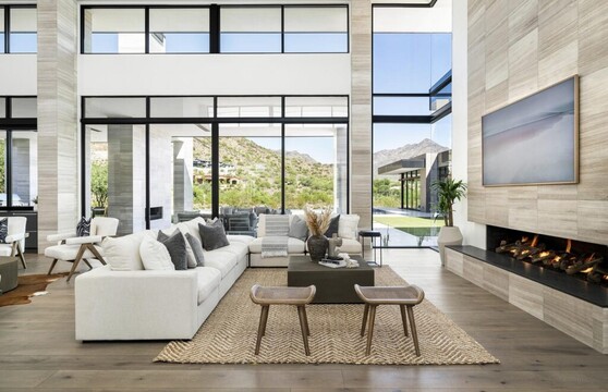 A contemporary living room is set against large glass sliding doors and windows that look out to the backyard and beyond to distant neighbors’ homes, and mountains beyond that.