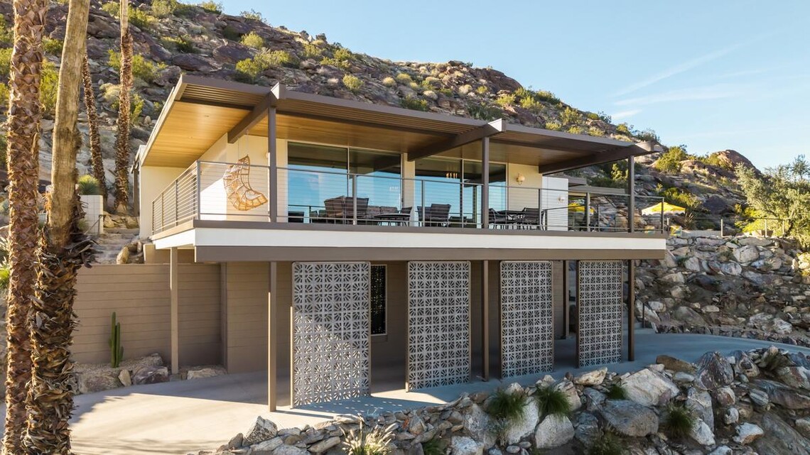 A modern home’s exterior is visible in front of a rocky desert mountain. The home has mid-century modern style, including a flat roof, large glass sliding doors on the upper level, and decorative breeze blocks on the lower level. 