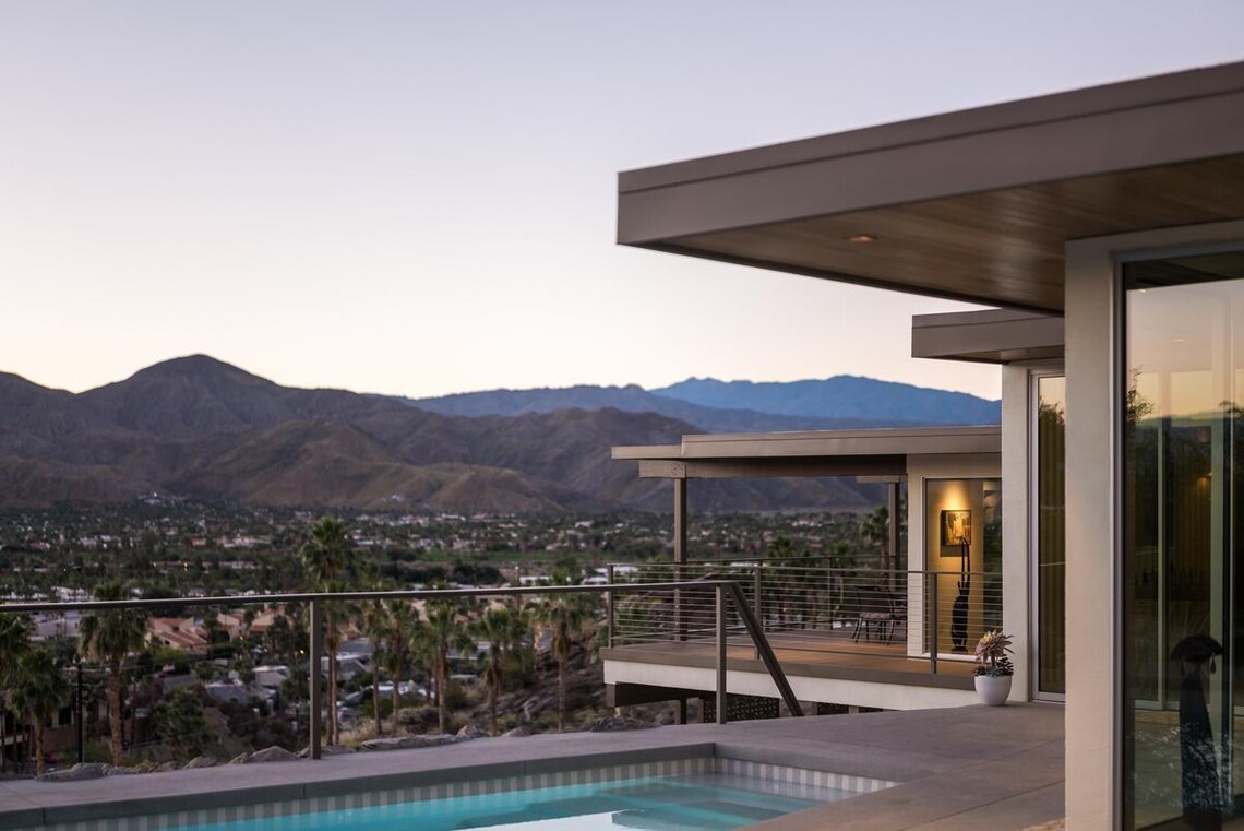 In the foreground, a home with glass walls and a flat roof are just barely visible to the right, along with a deck with a pool. The main view is of a town with palm trees and mountains in the distance.