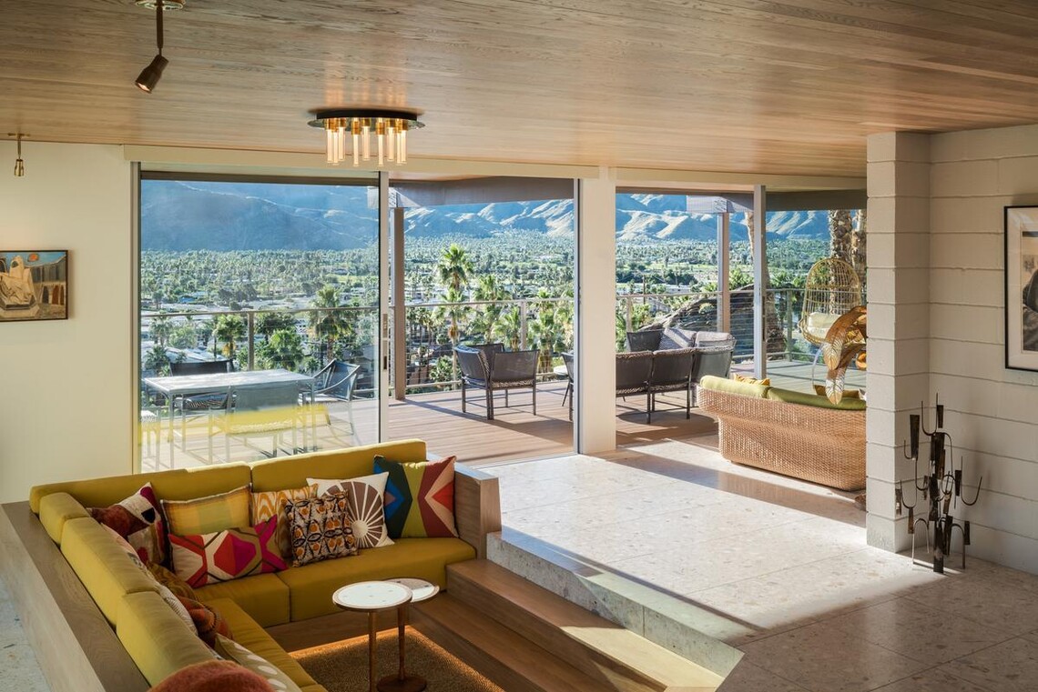 A conversation pit surrounded by a yellow sofa with colorful patterned throw pillows make up the living room in the foreground, and behind it, open sliding doors lead to the furnished deck that overlooks the Palm Springs below, and mountains beyond that.