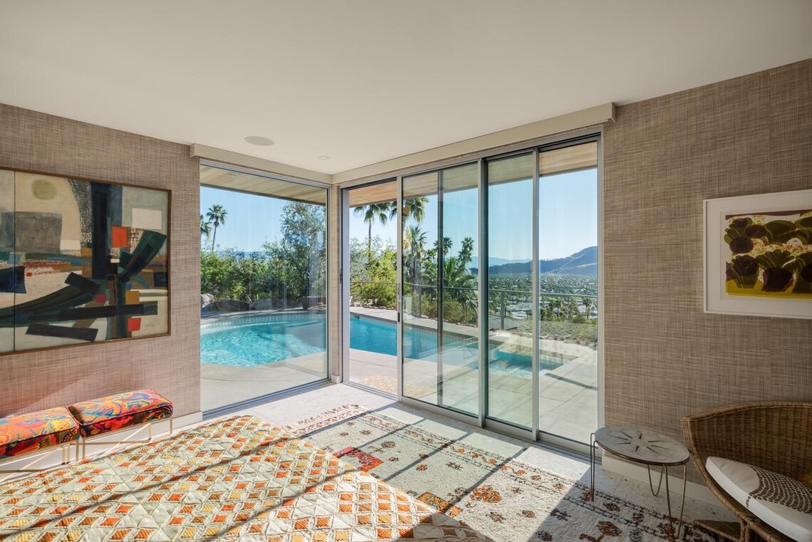 A multi-slide door is kitty-corner to a floor to ceiling window. The door is partially open, leading from a bedroom to a deck with a pool outside. 