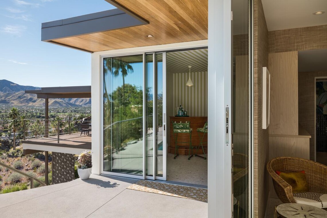 A home’s open sliding doors in the foreground show peaks at modern furnishings from a deck, and just beyond, another furnished deck is visible that overlooks desert landscaping. 