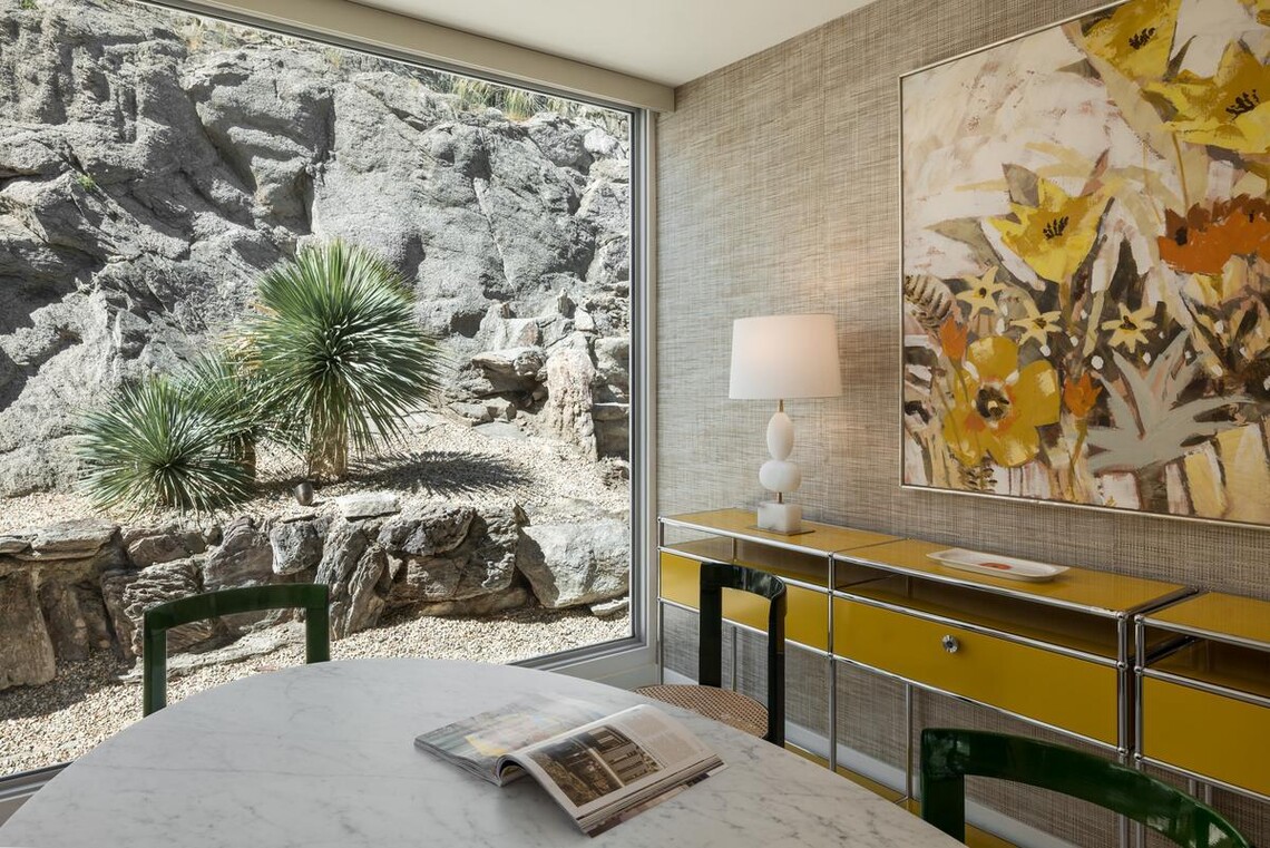 A yellow and chrome mid-century modern credenza is against a dining room wall with a large painting of yellow and orange flowers, and a dining room table in the foreground. To the left is a floor-to-ceiling window that displays the mountain the home is built into, and desert vegetation. 
