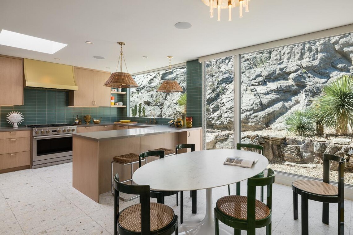 A dining room table and chairs are in front of a kitchen island, and both are in front of floor-to-ceiling windows that show the rocks and desert plants of the mountain that the home is built into. 