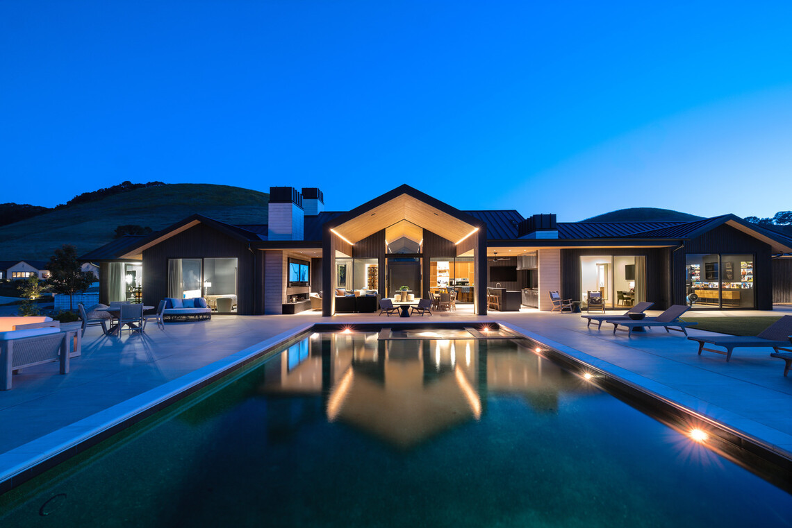 San Luis Obispo Vineyard Residence illuminated at dusk, with expansive glass openings overlooking a swimming pool and surrounding hills.
