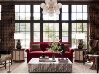 Elegant living room with floor-to-ceiling black-framed windows behind a red velvet sofa, flanked by matching lamps on round side tables, a marble coffee table, and a striped accent chair.