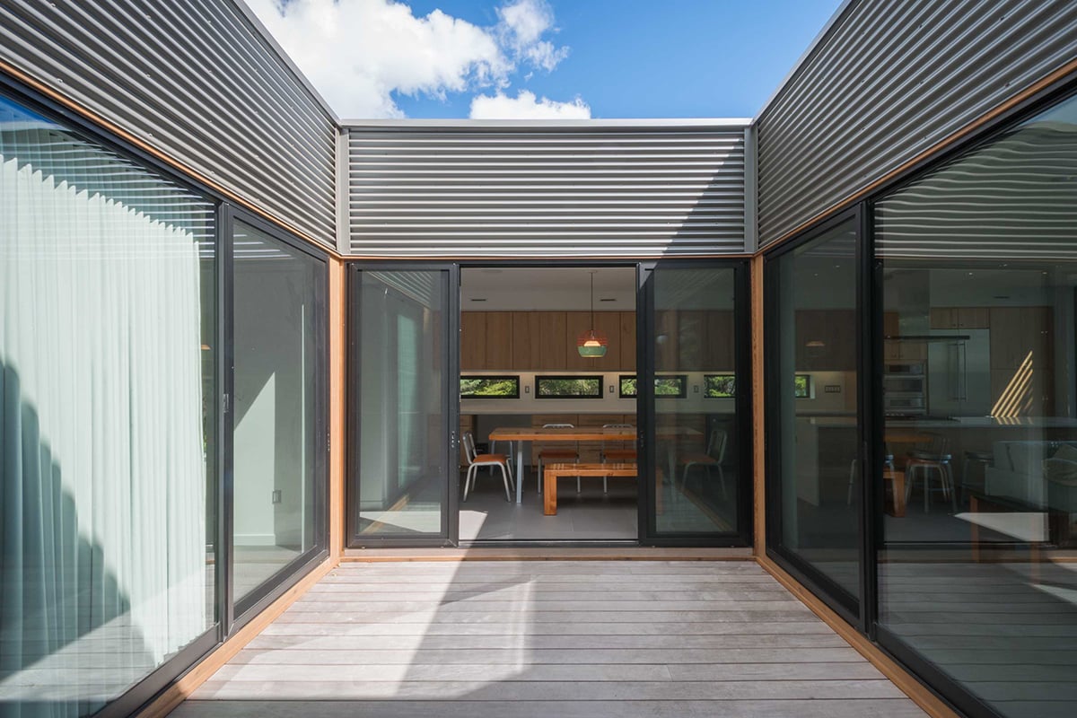 A view of an outdoor corridor leading into the dining area through open, multi-slide doors surrounded by large panels of glass.
