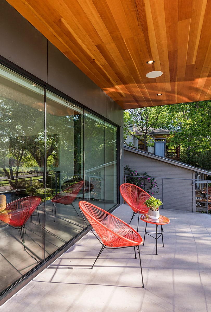 The front porch has two orange chairs and a small table with their backs to a large butt-glazed window wall that shows minimum interruption between panes.