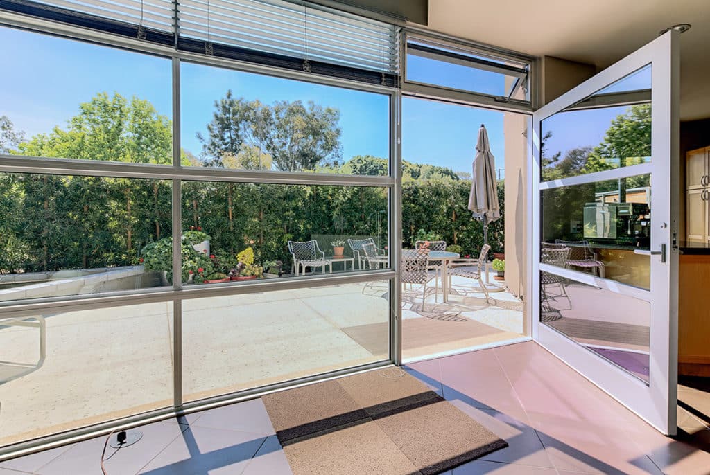 The massive hinged-door and accompanying wall of windows opens to a back patio surrounded by shrubbery.