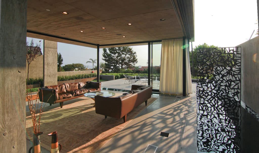 The stunning sunroom with two brown leather couches around a glass table is surrounded on three sides by immense moving panels of glass.