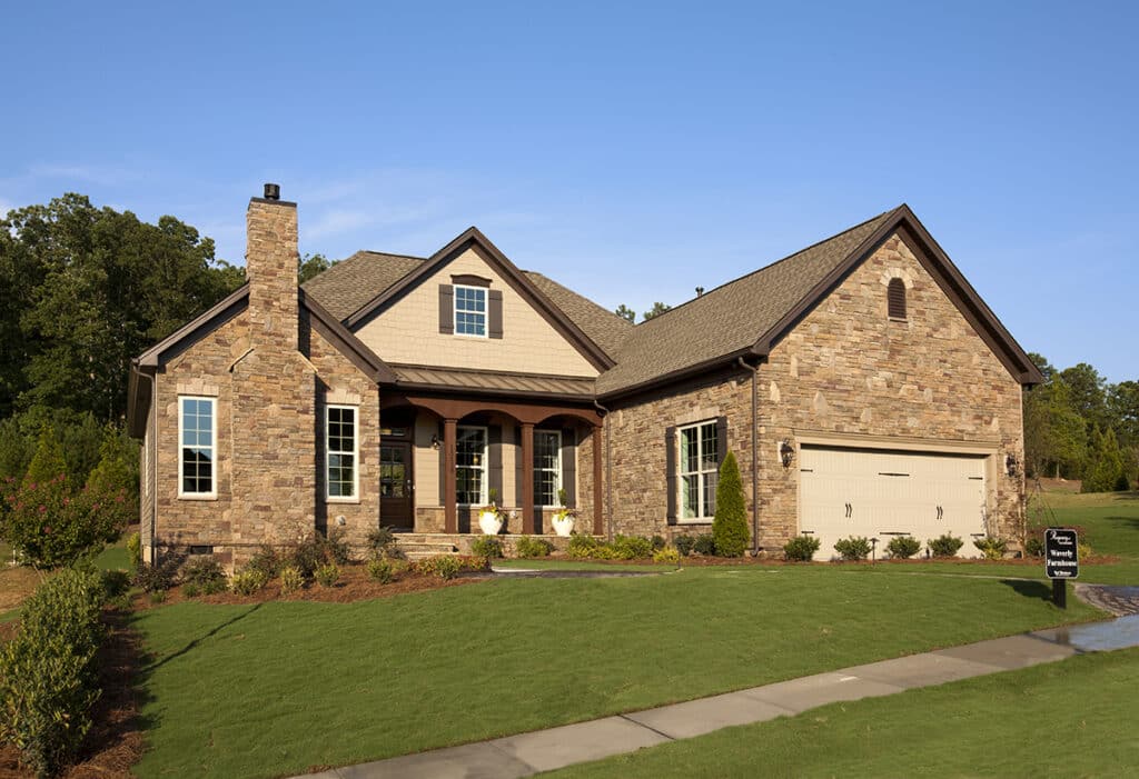A view of the outside of a one-story house with warm brown brick framed by bright green trees and grass.