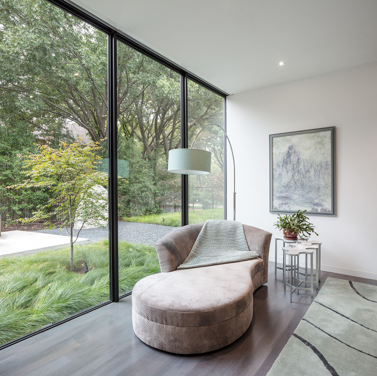 The window wall next to a long lounge chair frames views of grass and trees.