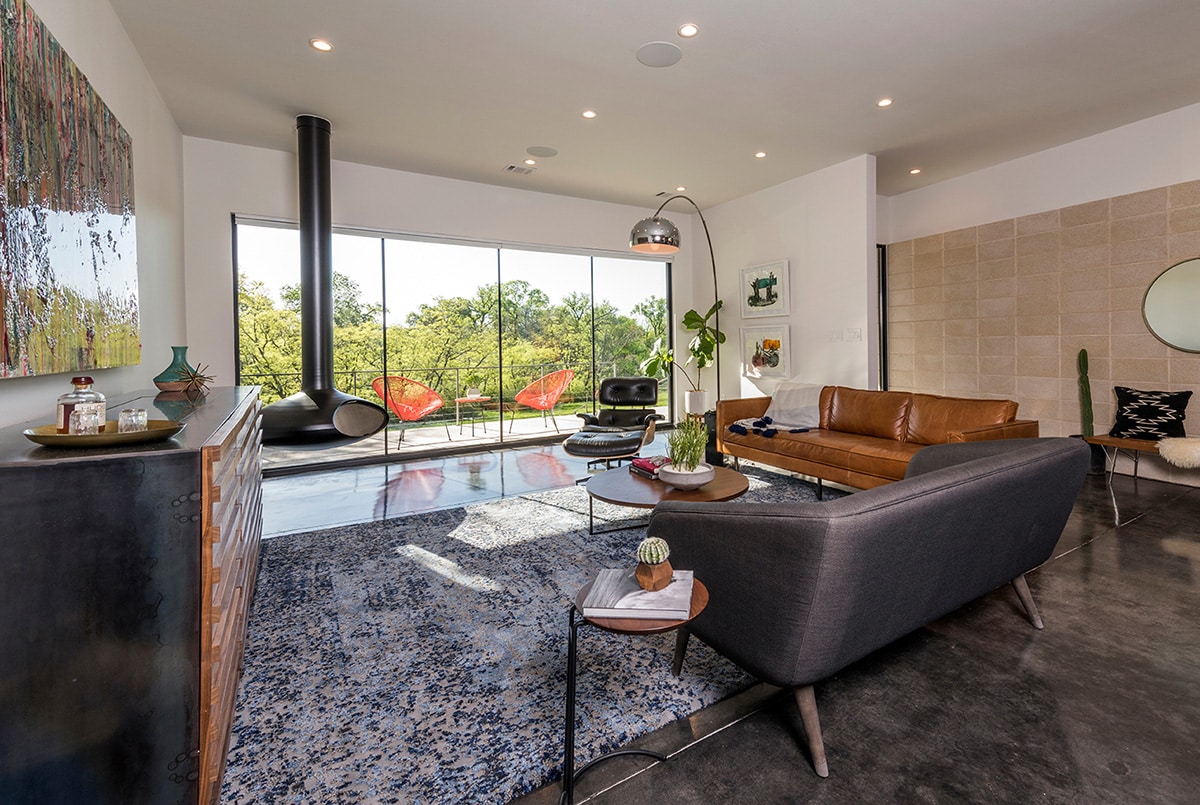 A modern living room looks out a large window wall to the front porch with a view of trees from the park.