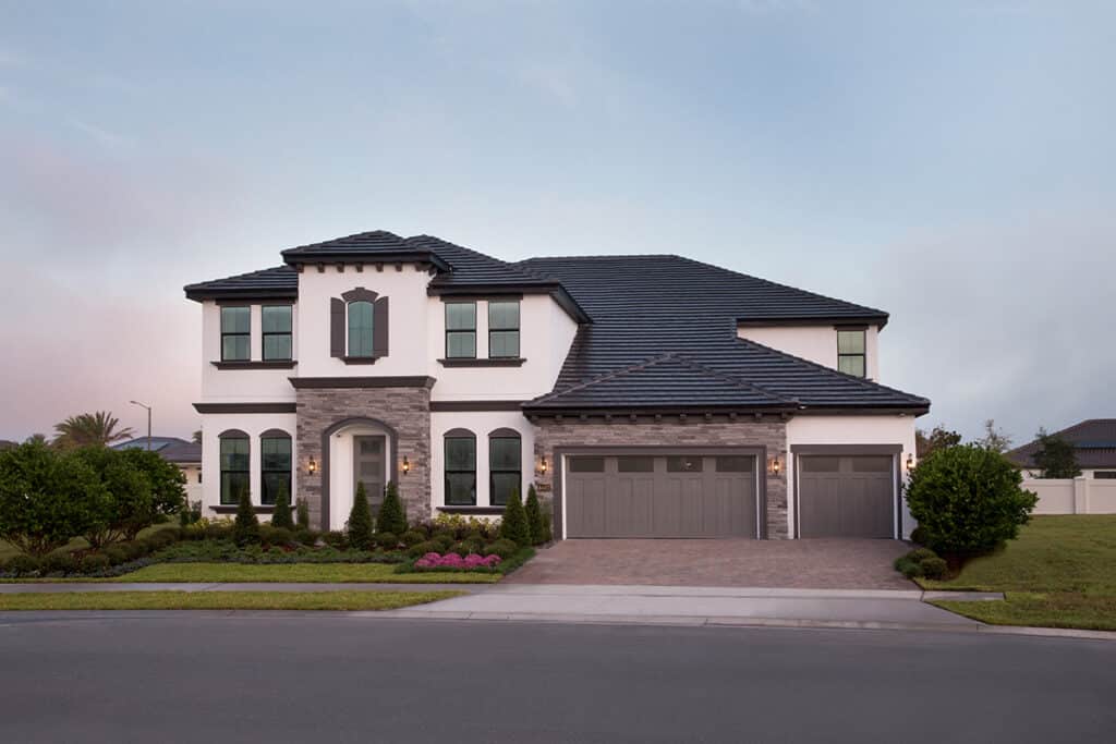 A two-story home with a white and brown exterior and a dark brown roof is framed by a lush green yard and sunset sky.