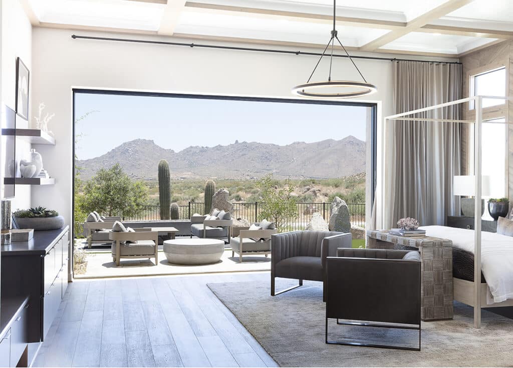 A tall multi-slide glass door is fully open, allowing a bedroom to transition to an outdoor seating area and view of a mountain landscape.