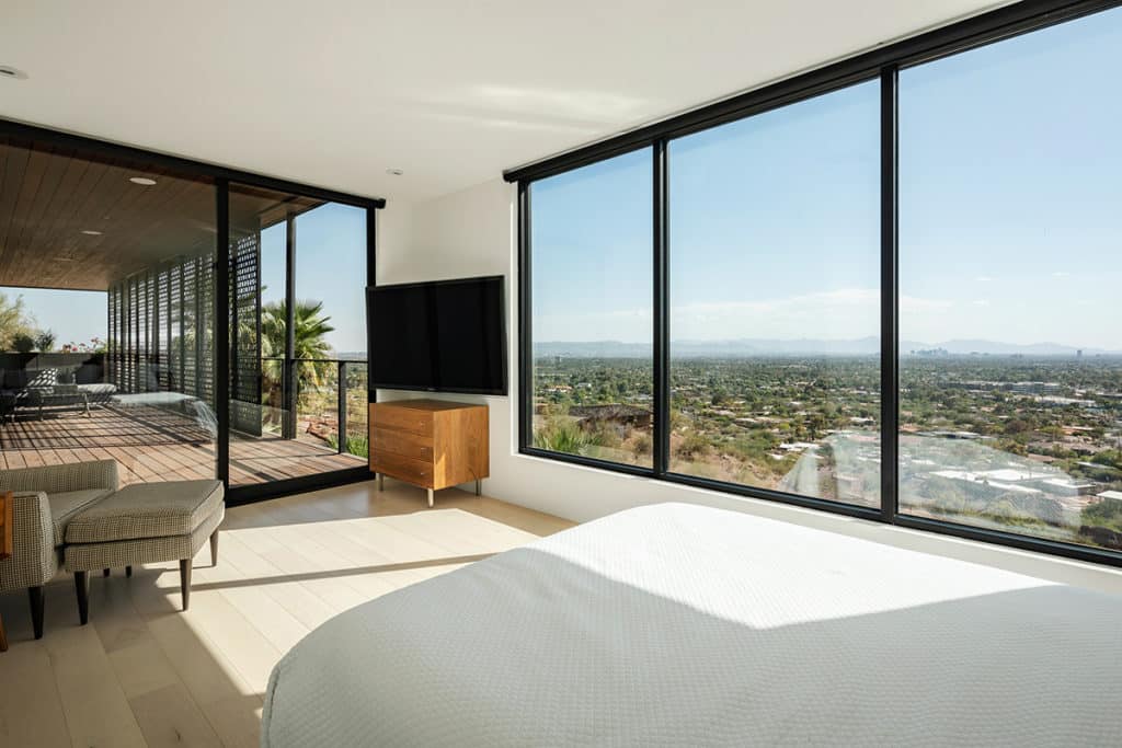 The fixed windows in this bedroom provides a view of valley residences while a large multi-slide door connects to an outdoor seating area.