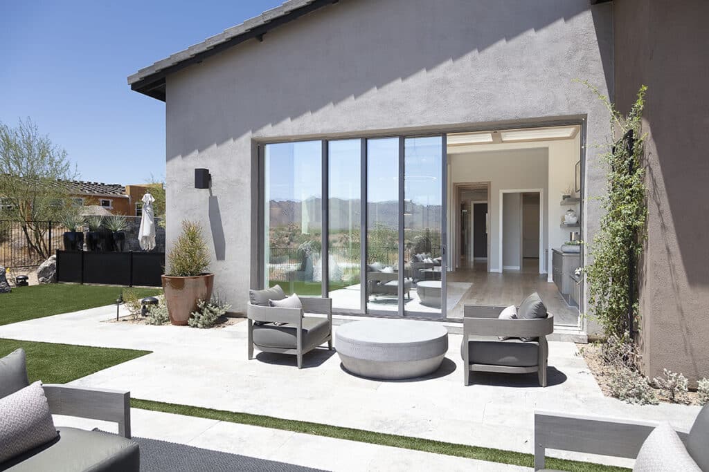 An outdoor seating area leads to an indoor bedroom through a partially open multi-slide glass door.