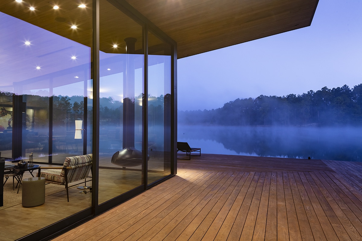 A mist covers the lake at sunrise; a glass building and deck on the shore.